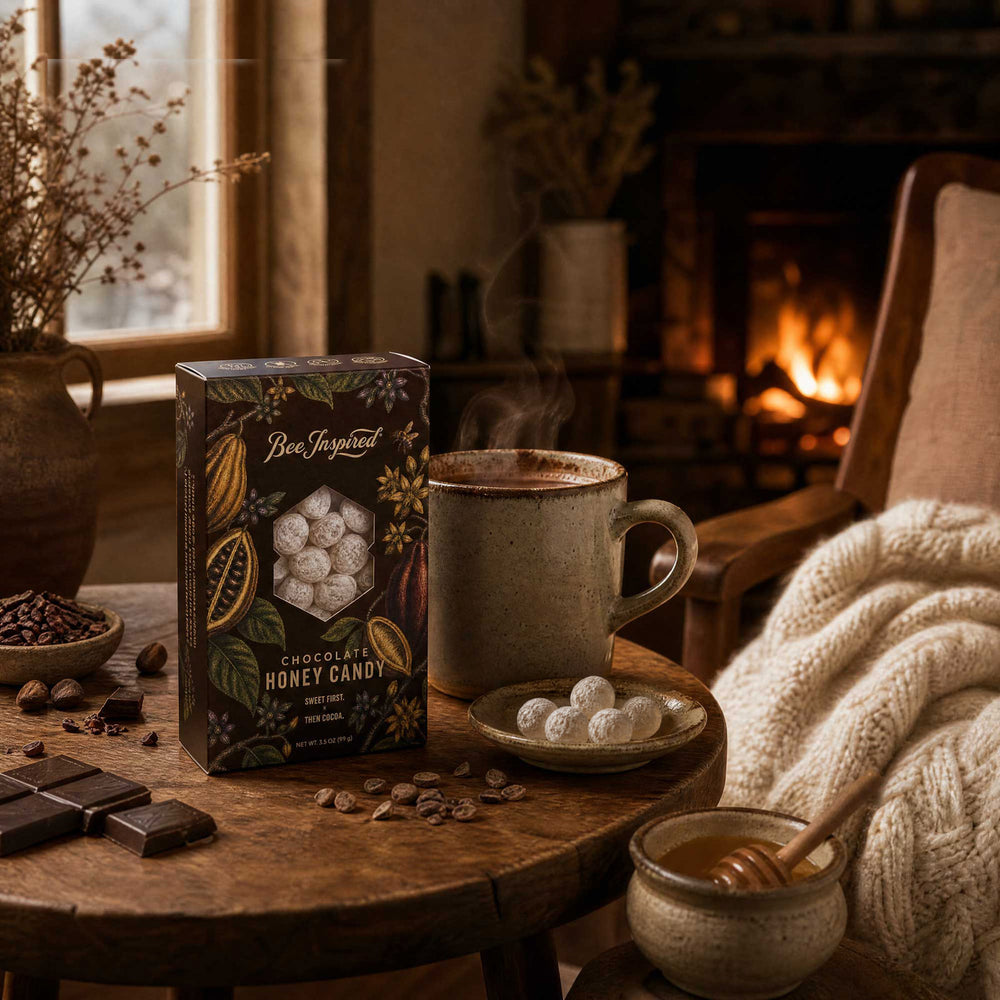 A box of Chocolate Honey Candy stands on a rustic wooden table beside a steaming mug, small plates with candied fruit, chocolate pieces, and a cozy fireplace scene in the background.