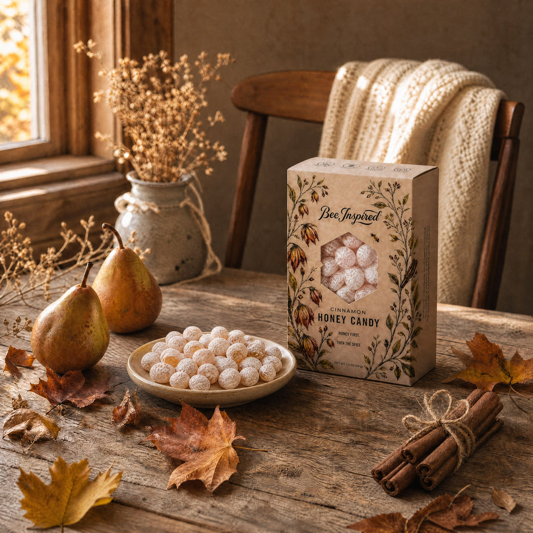 A styled tabletop scene showing a box of bee-inspired honey candy next to a bowl of powdered sugar-coated honey candies, with pears, autumn leaves, cinnamon sticks, and a knitted blanket in a warm rustic room.