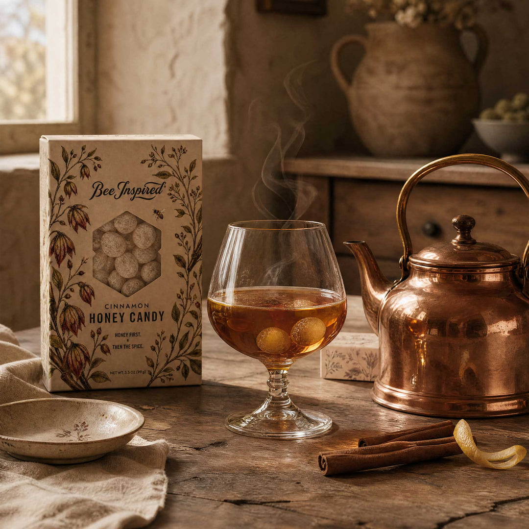 A box of Bee Inspired Cinnamon Honey Candy sits on a rustic wooden table next to a glass of amber-colored honey, a copper teapot, cinnamon sticks, and a small dish, with steam rising from the glass.