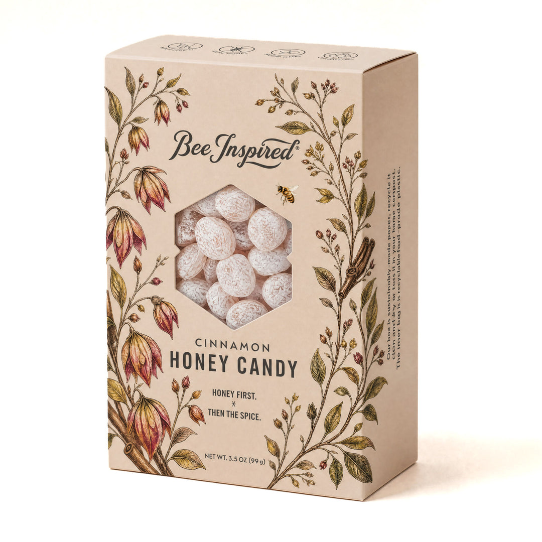 A rectangular beige box of Bee Inspired Cinnamon Honey Candy, featuring a hexagonal window that reveals round, frosted white candies inside. The packaging is decorated with vintage-style botanical illustrations of flowers and leaves in green, brown, and red, with the brand name 'Bee Inspired' printed at the top.