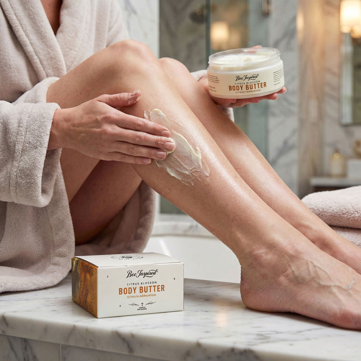 Person applying body butter to their legs with a jar and box of the product in a bathroom setting.