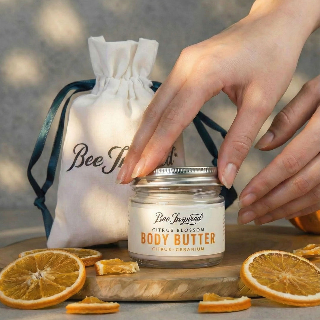 Hand holding a jar of 'Bee Inspired' body butter with oranges and a drawstring bag in the background.