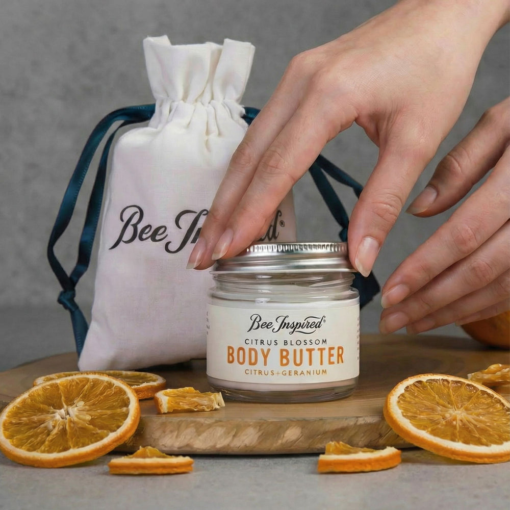 Jar of 'Bee Inspired' body butter with a hand reaching for it, surrounded by orange slices on a wooden board.
