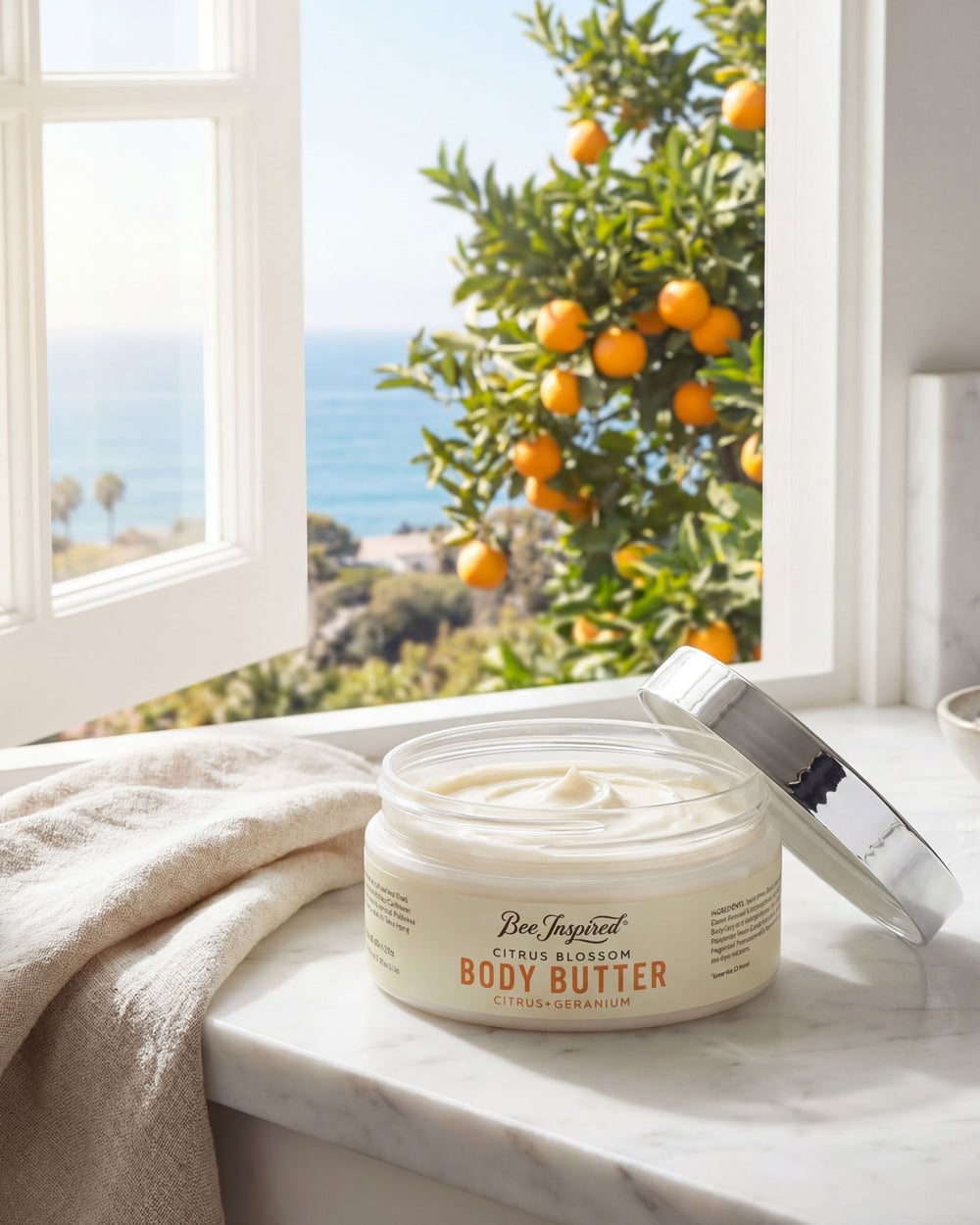Jar of body butter on a windowsill with an orange tree and ocean view outside.