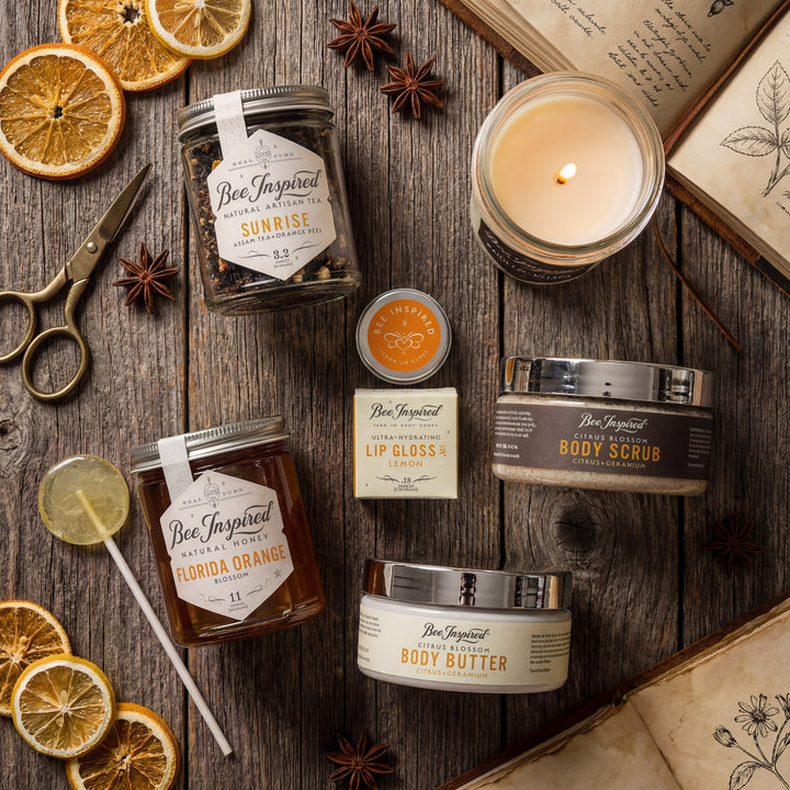 Collection of Bee-Inspired products including candles, lip balms, and body scrubs on a wooden surface with dried oranges and star anise.
