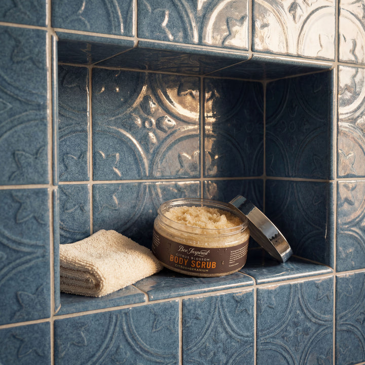 Jar of body scrub on a tiled shelf with a towel and loofah.