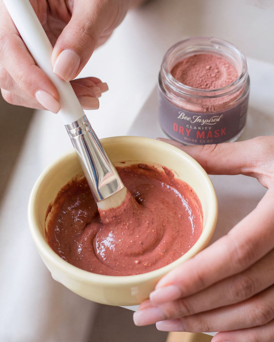 Person mixing a pink clay mask in a bowl with a brush, next to a jar of the same mask.
