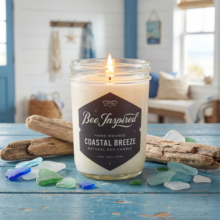 Candle labeled 'Bee Inspired' with 'Coastal Breeze' scent on a table with driftwood and sea glass.