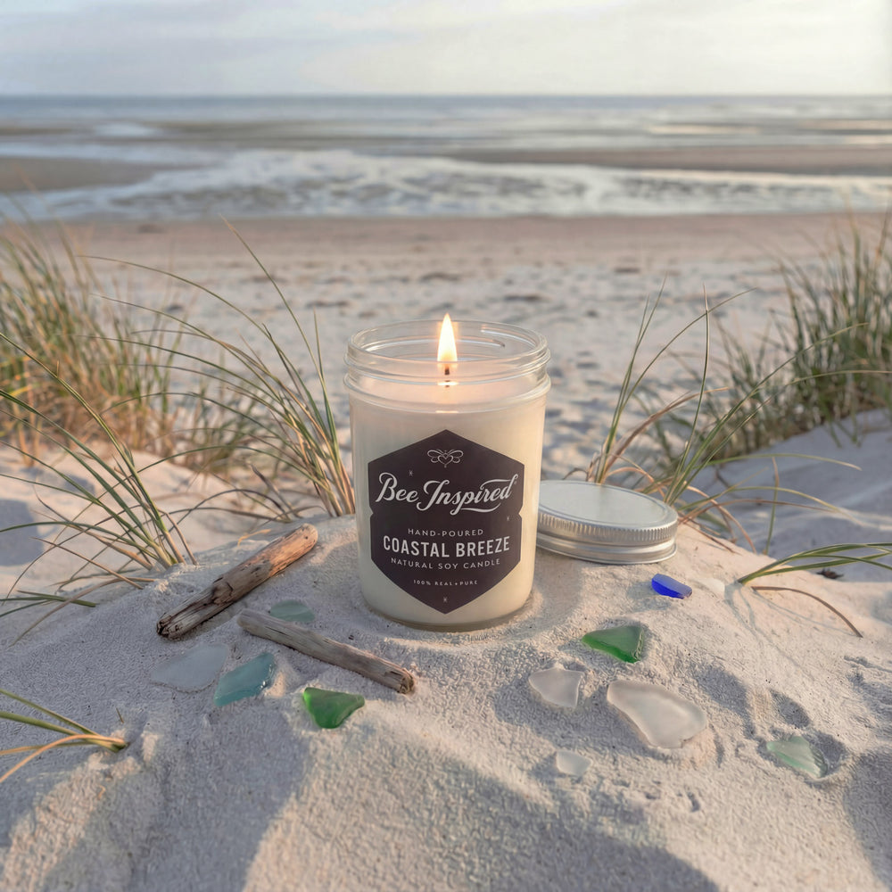 Candle labeled 'Bee Inspired Coastal Breeze' on a sandy beach with grass and beach glass.