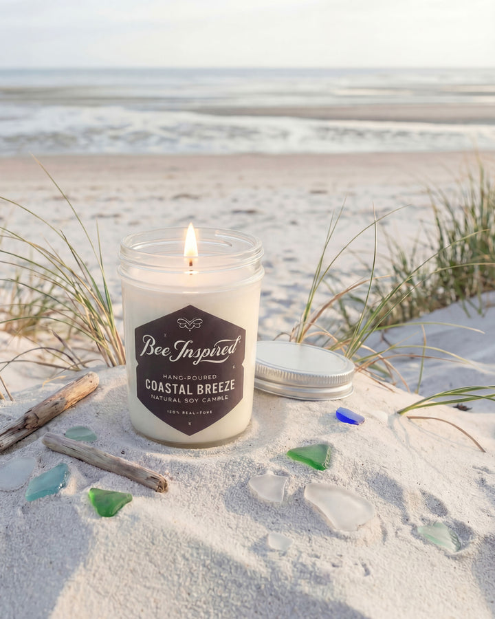 Candle labeled 'Bee Inspired Coastal Breeze' on a sandy beach with ocean in the background