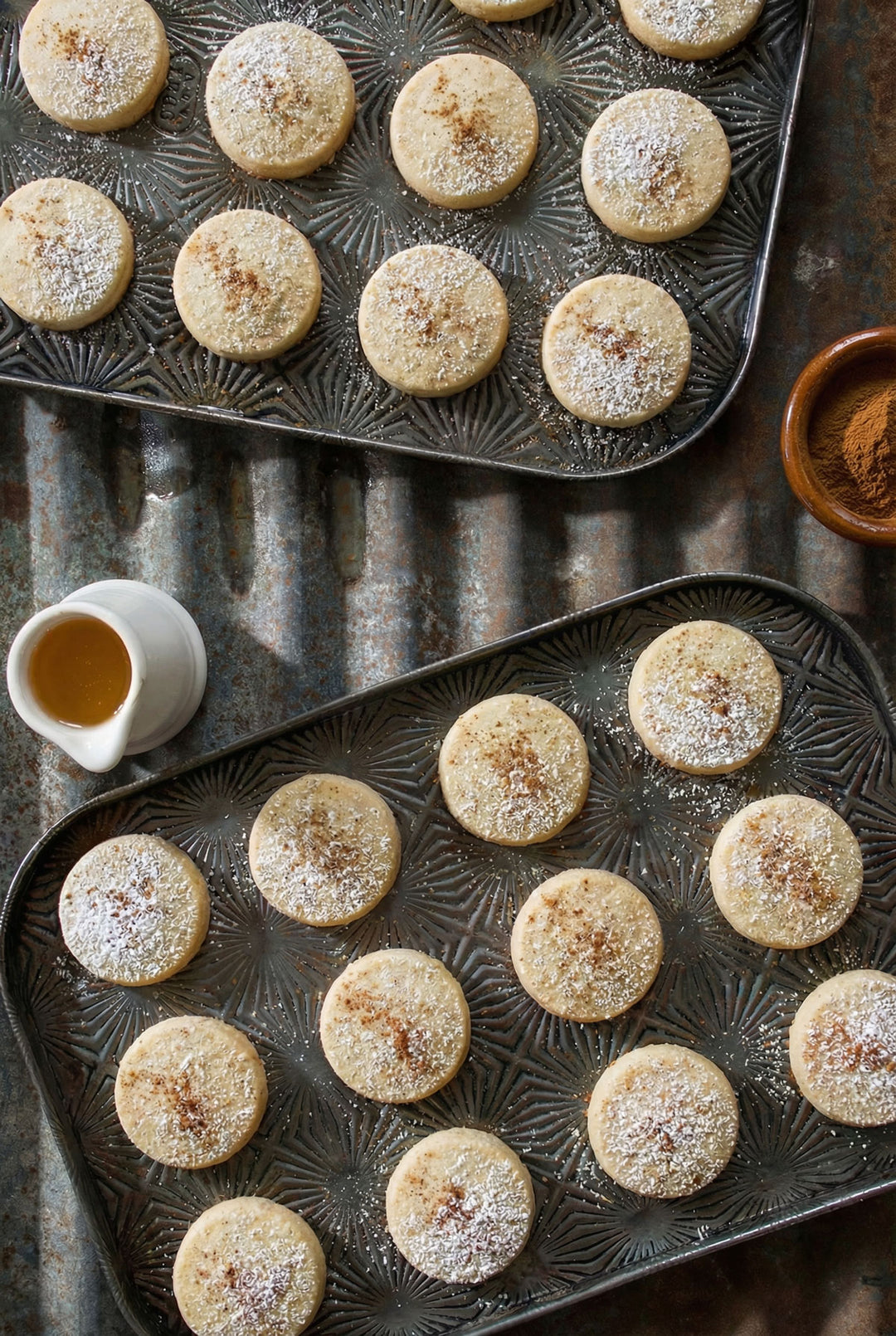 Baked cookies on a metal baking tray with a small bowl of cinnamon and a pitcher of honey