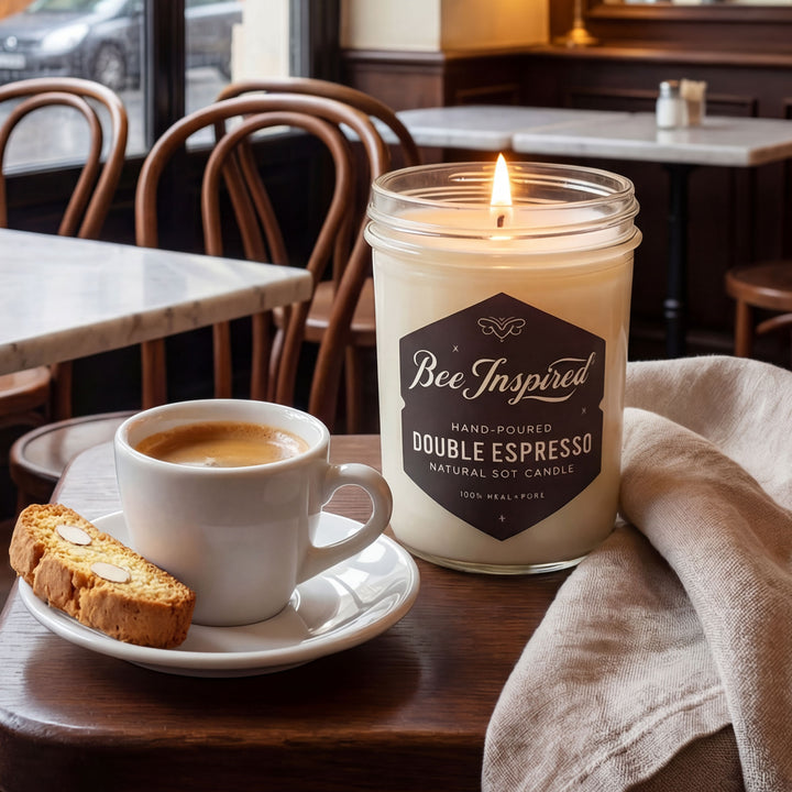 Candle labeled 'Bee Inspired Double Espresso' with a cup of coffee and biscotti on a table in a cafe.