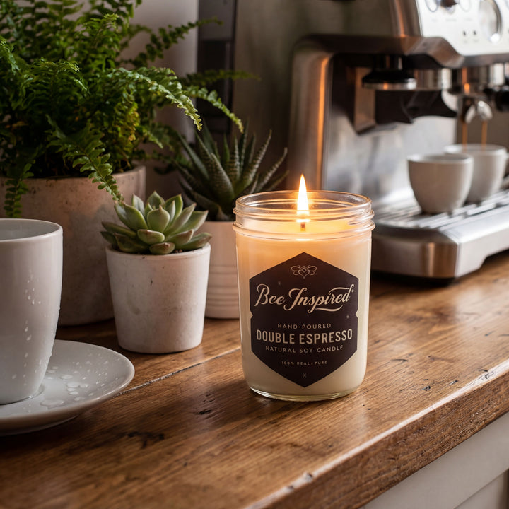 Candle labeled 'Bee Inspired Double Espresso' on a wooden surface with coffee cups and plants in the background.
