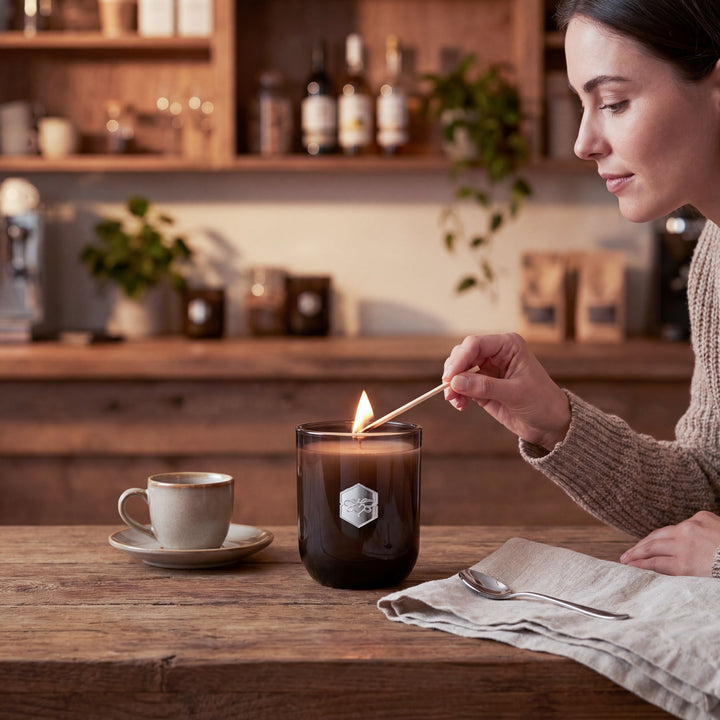 Woman lighting a candle in a kitchen setting with a cup of coffee and a spoon on a wooden table.