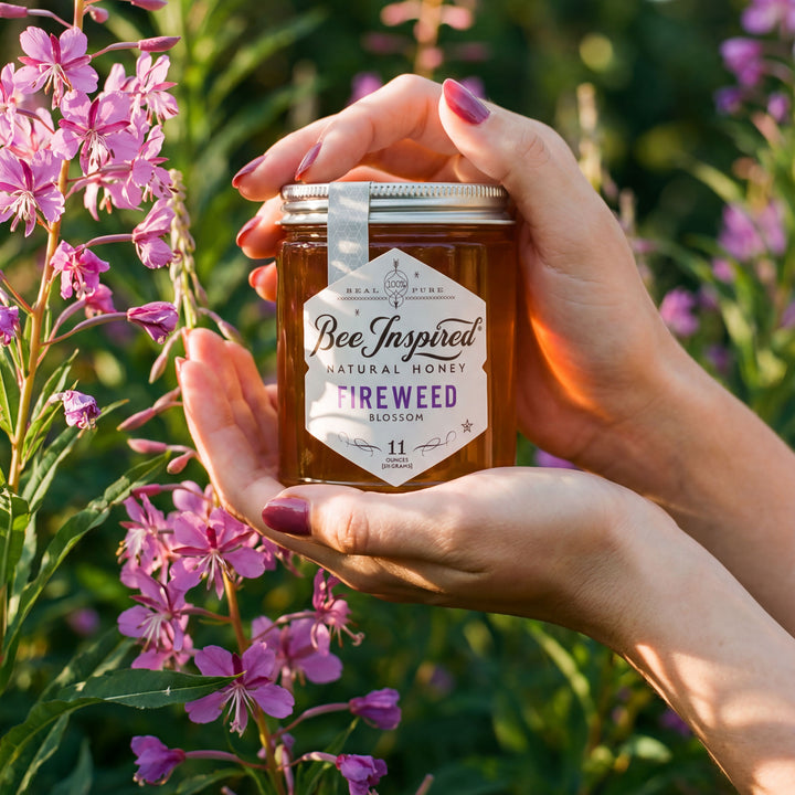 Hand holding a jar of 'Bee Inspired' natural honey with fireweed flowers in the background