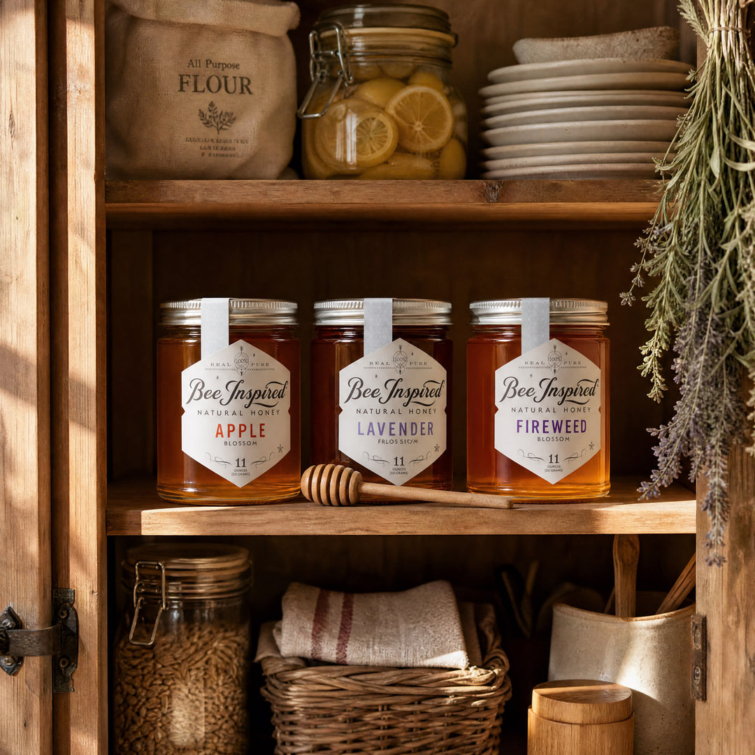 Three glass jars of honey displayed on rustic wooden shelves inside a cabinet, surrounded by natural kitchen and beekeeping props including a honey dipper, plates, flour sack, and lavender sprigs. The labels read Bee Inspired Natural Honey in Apple, Lavender, and Fireweed flavors.