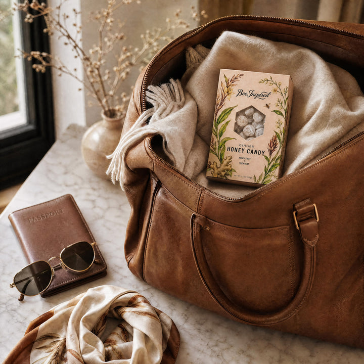 A brown leather duffel bag sits open on a marble surface, filled with a cream knit blanket and a box of Bee Sweetheart honey candy. A brown passport holder, round sunglasses, and a cream scarf are placed beside it near a window with dried flowers in a vase.
