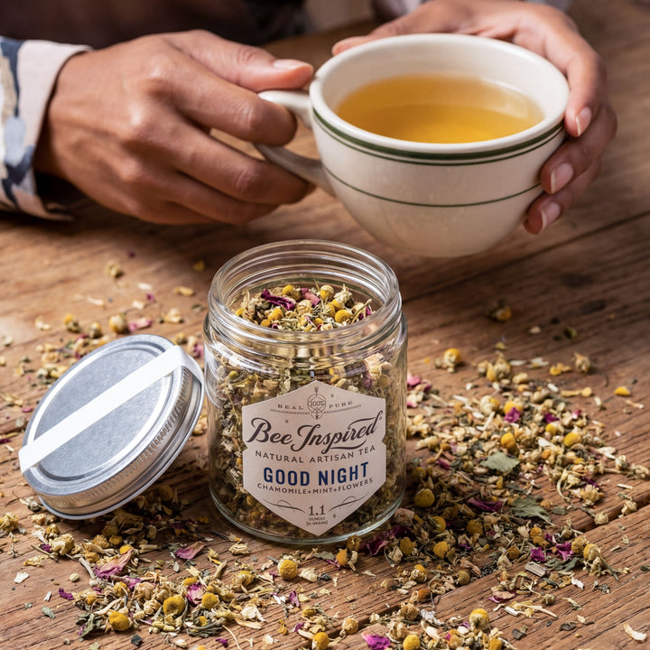 Person holding a cup of tea with a jar of 'Good Night' herbal blend on a wooden surface.