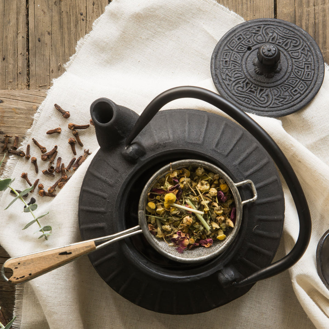 Black teapot with a strainer containing dried herbs on a wooden surface.