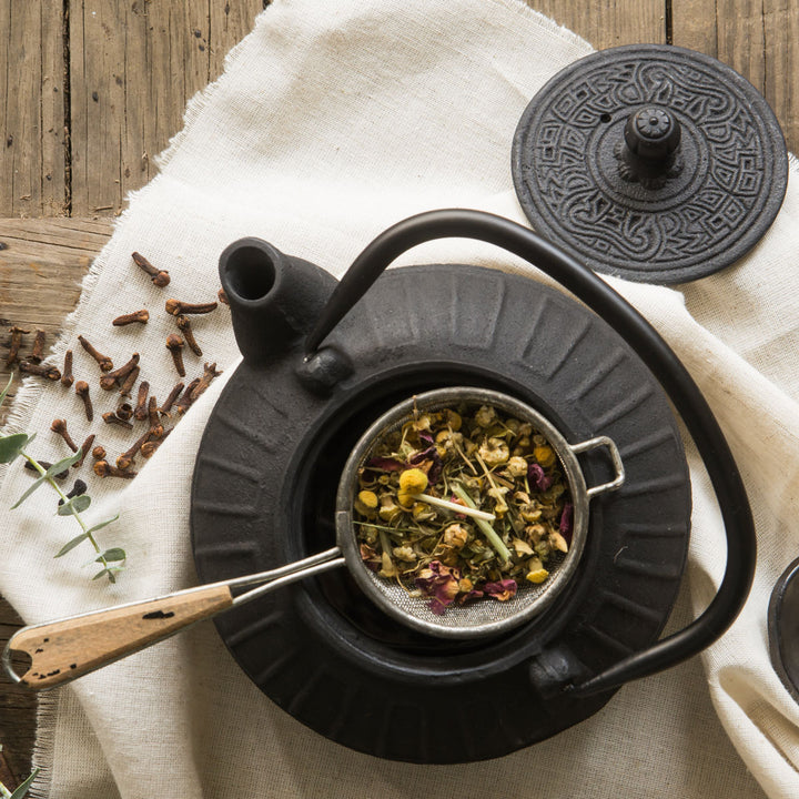 Black teapot with a strainer containing dried herbs on a wooden surface.