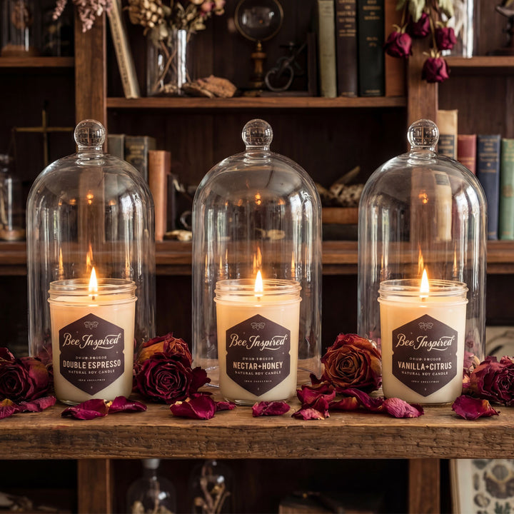 Three lit candles in glass domes on a wooden surface with flowers and books in the background