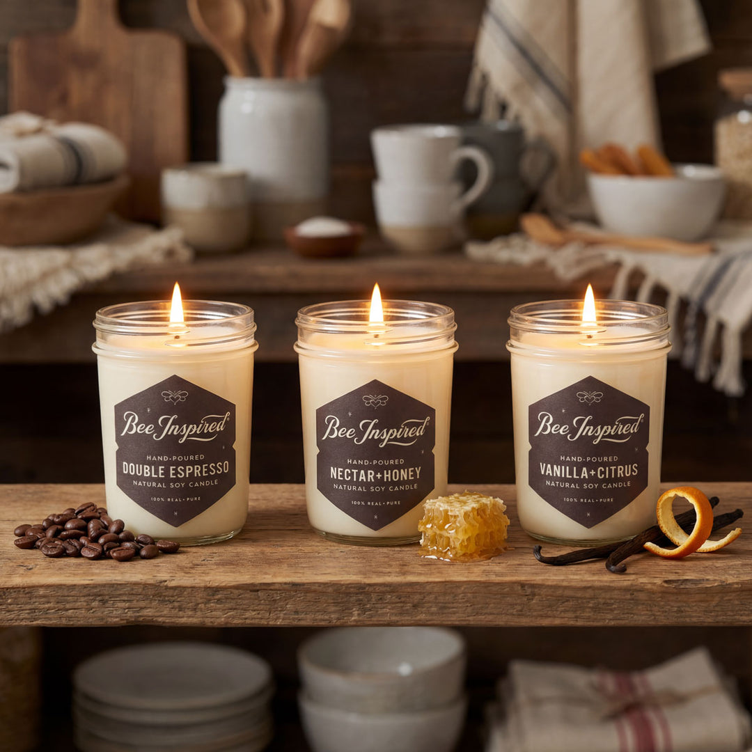 Three lit candles labeled 'Bee Inspired' on a wooden surface with coffee beans, honeycomb, and vanilla pods.
