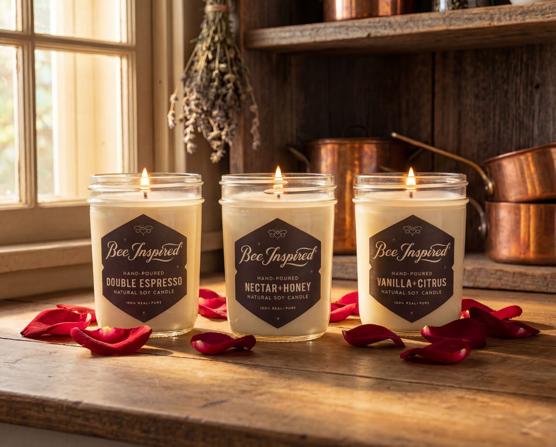 Three lit candles labeled 'Bee Inspired' on a wooden surface with red flower petals.