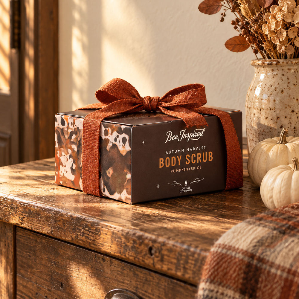 A boxed body scrub product sits on a rustic wooden table in a warm, styled indoor setting. The box is dark brown with a patterned side panel and tied with a rust-brown ribbon bow.