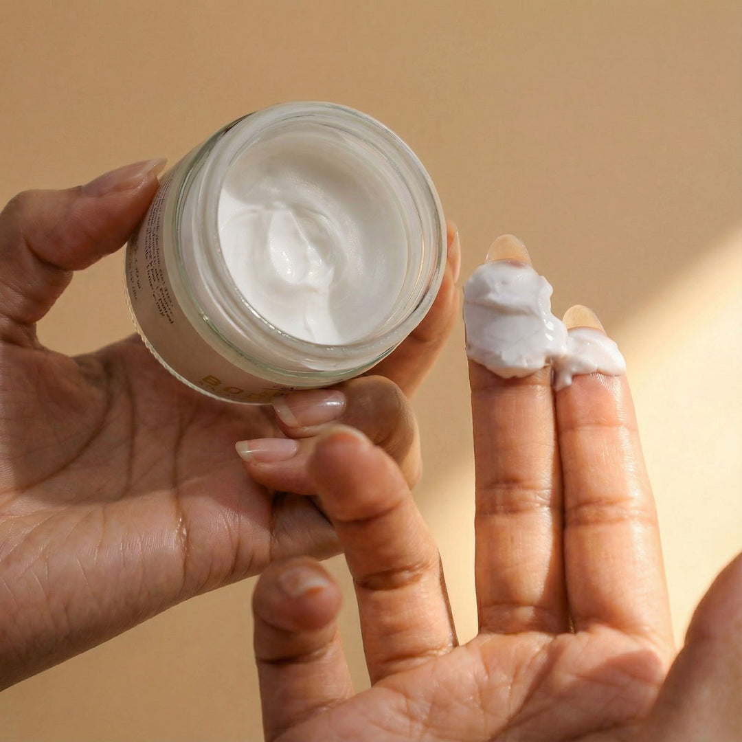 Person holding a jar of cream with a hand applying some to the fingers against a beige background