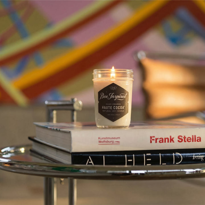 Candle labeled 'Bee Inspired' on top of books with a colorful abstract painting in the background