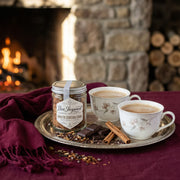 Two cups of hot chocolate with a jar of cocoa mix on a tray in front of a fireplace.