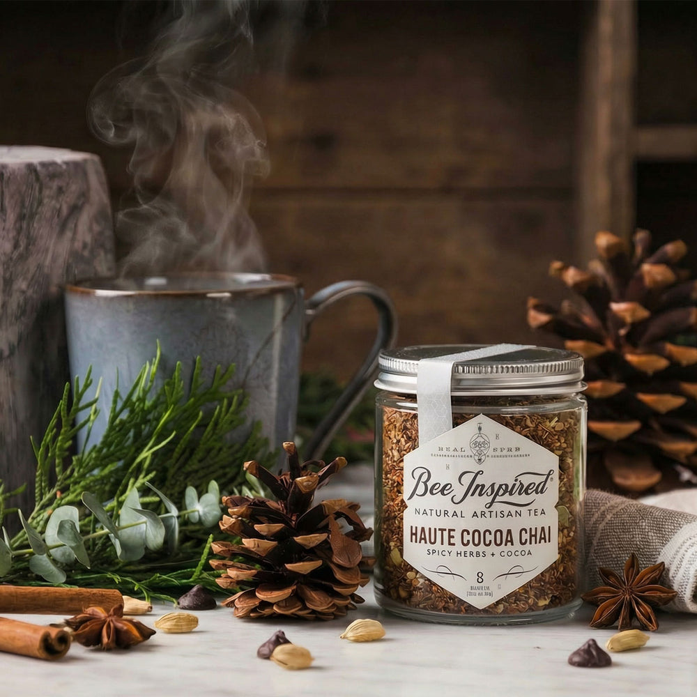 Jar of 'Bee Inspired' haute cocoa chai tea with a steaming mug and pinecones on a wooden surface.