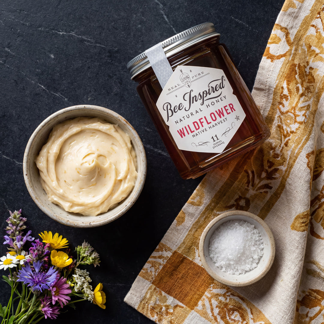 Jar of Bee Inspired Wildflower Honey with a bowl of honey and flowers on a dark surface.