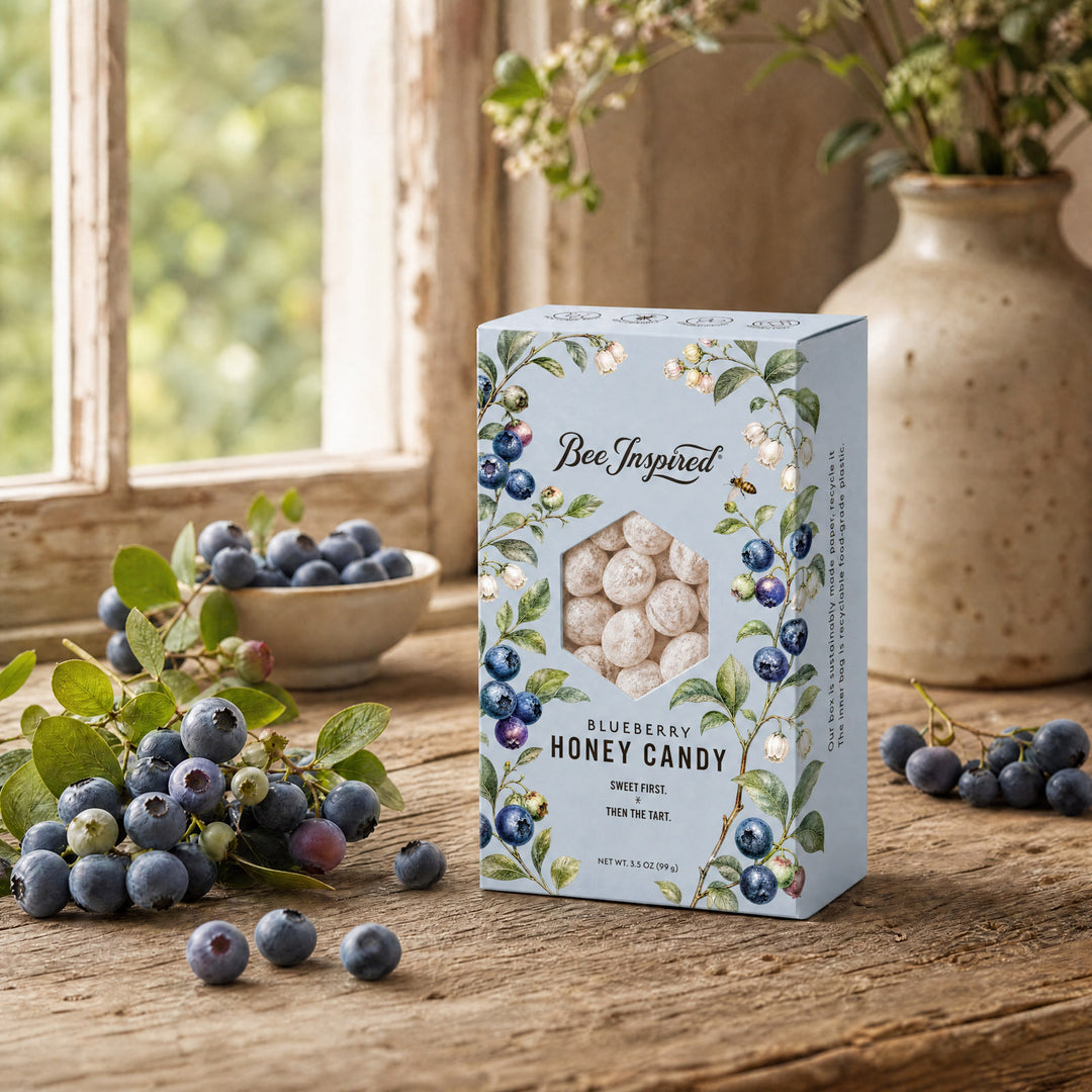 A box of Bee Inspired Blueberry Honey Candy sits on a rustic wooden table, surrounded by fresh blueberries, a small bowl, and a ceramic vase in a softly lit room.