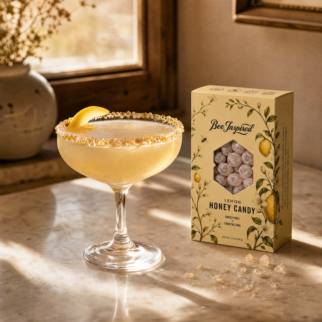 A boxed box of Bee Inspired Lemon Honey Candy sits next to a coupe glass filled with a pale yellow cocktail rimmed with crushed candy, on a sunlit stone surface.