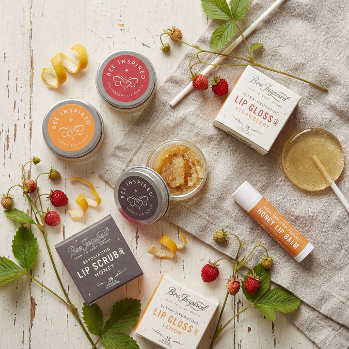 Collection of lip care products including lip gloss, lip balm, and scrub on a wooden surface with strawberries and leaves.