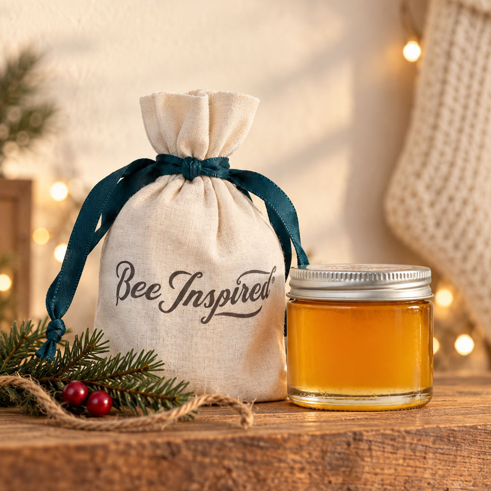 A small beige fabric drawstring bag with a dark teal ribbon and the words 'Bee Inspired' printed on the front, placed beside a small glass jar filled with golden honey on a wooden surface. The scene is styled with evergreen branches, red berries, and warm string lights in the background.