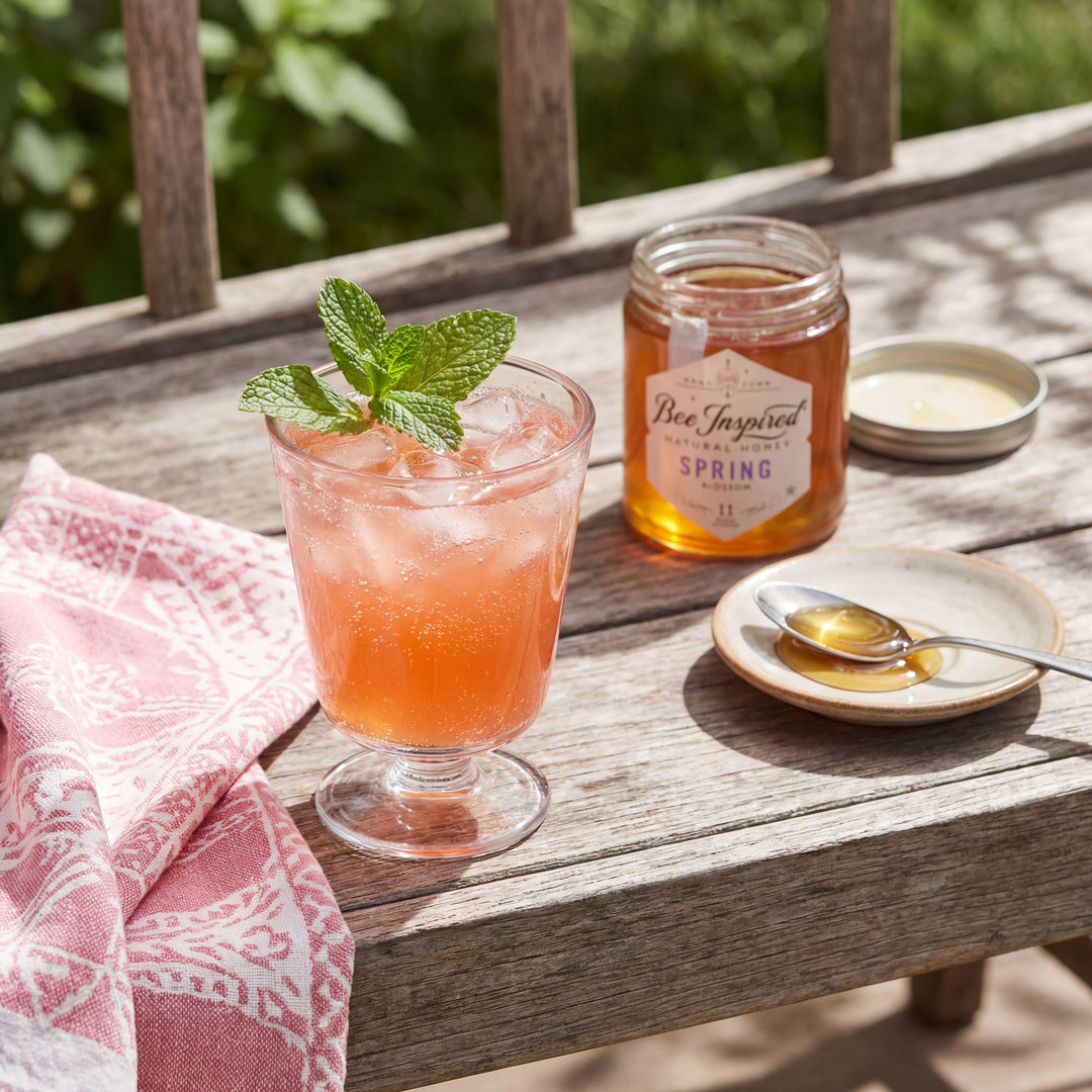 Glass of strawberry honey lemonaide iced drink with a mint leaf on a wooden table, next to a jar labeled 'Bee Inspired' and a spoon with honey.