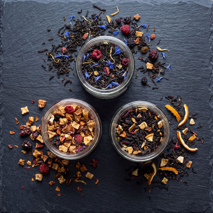 Three glass jars filled with assorted dried tea leaves on a dark slate surface.