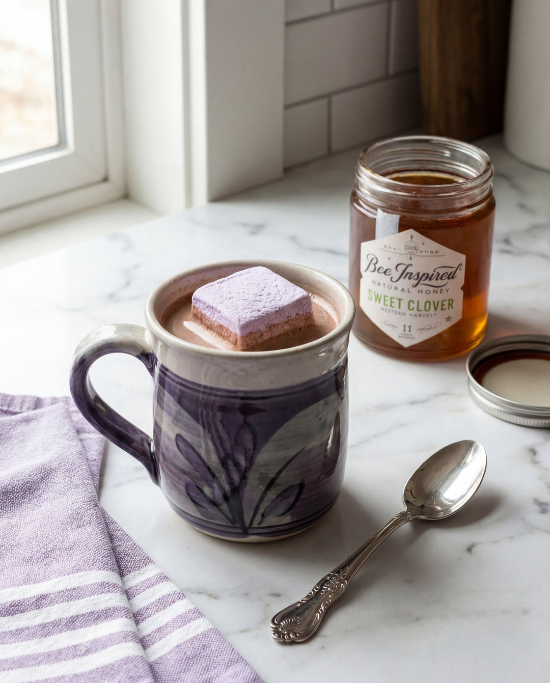 Mug of hot chocolate with marshmallows, jar of honey, and spoon on a marble surface.