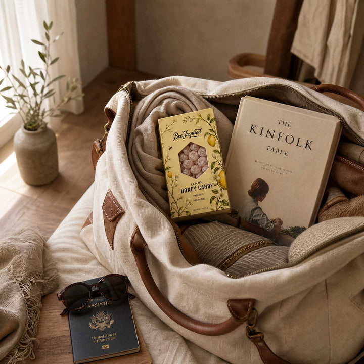 A large cream canvas duffel bag with brown leather handles sits open on a wooden floor, styled with a folded knit blanket, a yellow honey candy box, a book titled The Kinfolk Table, sunglasses, and a passport. A small vase with greenery is visible in the background near a window.