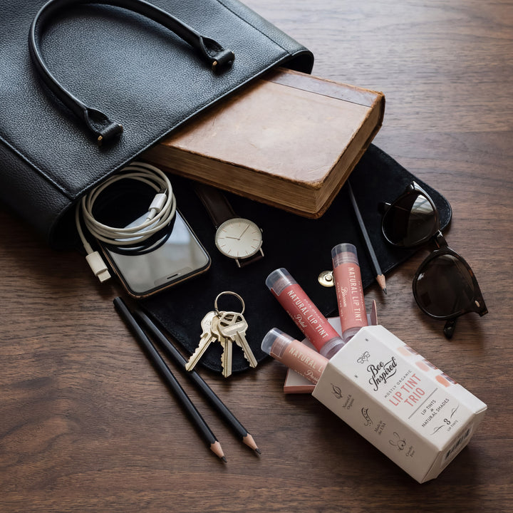 Black handbag open on a wooden surface with various items including lip products, pencils, and sunglasses.