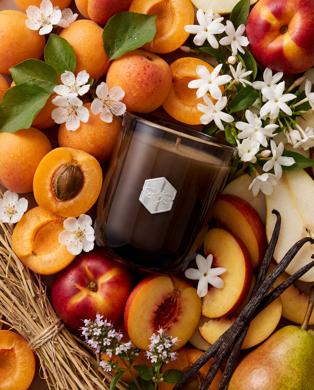 Candle surrounded by apricots, peaches, pears, and flowers on a woven mat.
