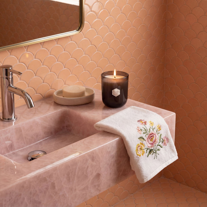 Bathroom sink with faucet, soap, candle, and embroidered towel on a tiled wall.