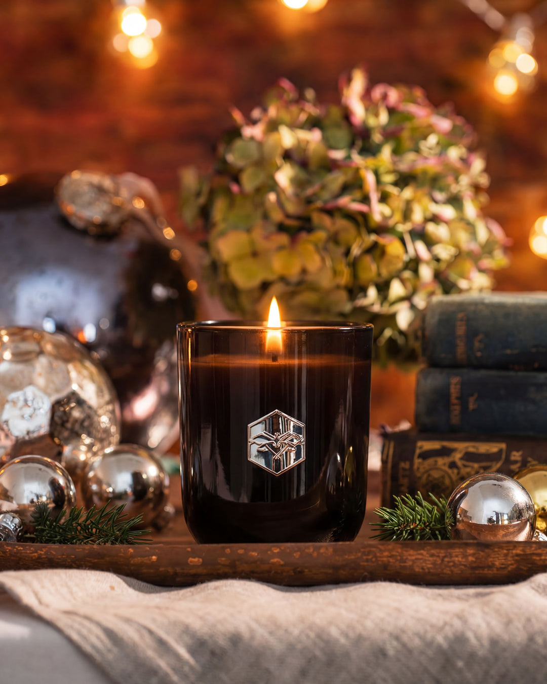 Candle in a black glass holder with a logo on a wooden tray, surrounded by decorative items.