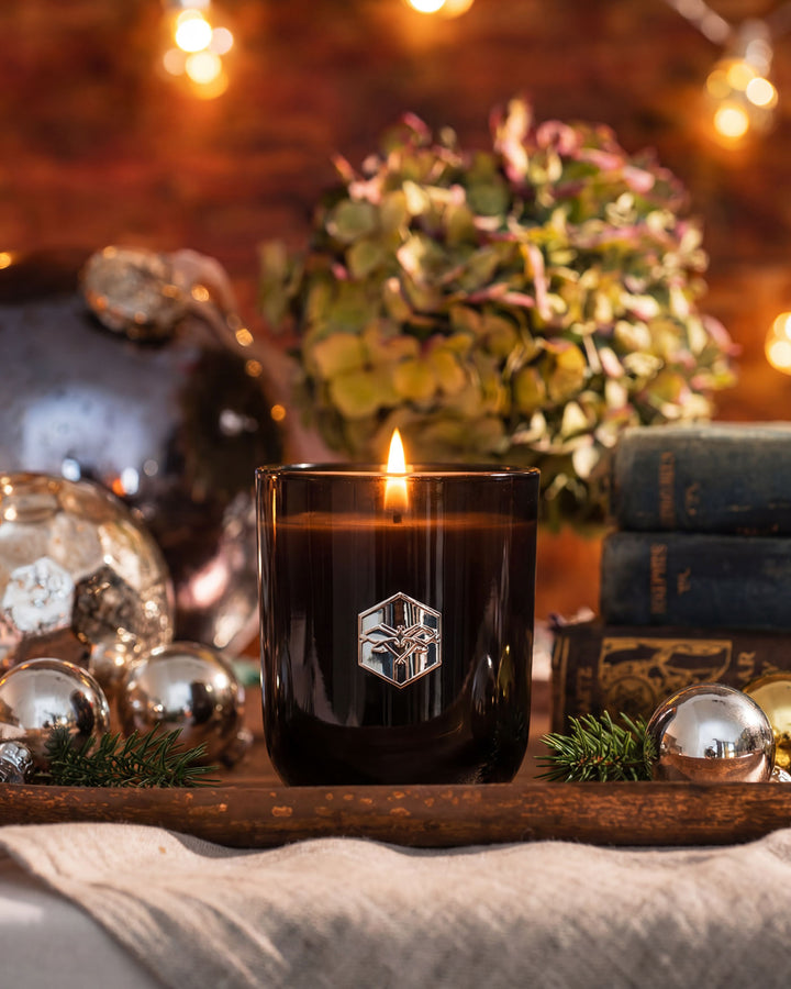 Candle in a black glass holder with a logo on a wooden tray, surrounded by decorative items.