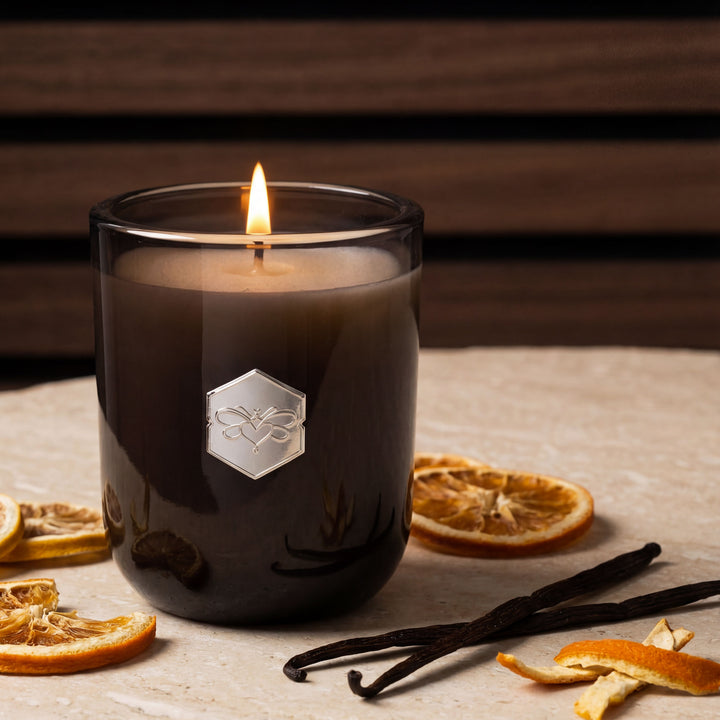 Candle in a glass jar with a logo, surrounded by vanilla beans and dried oranges on a wooden surface.