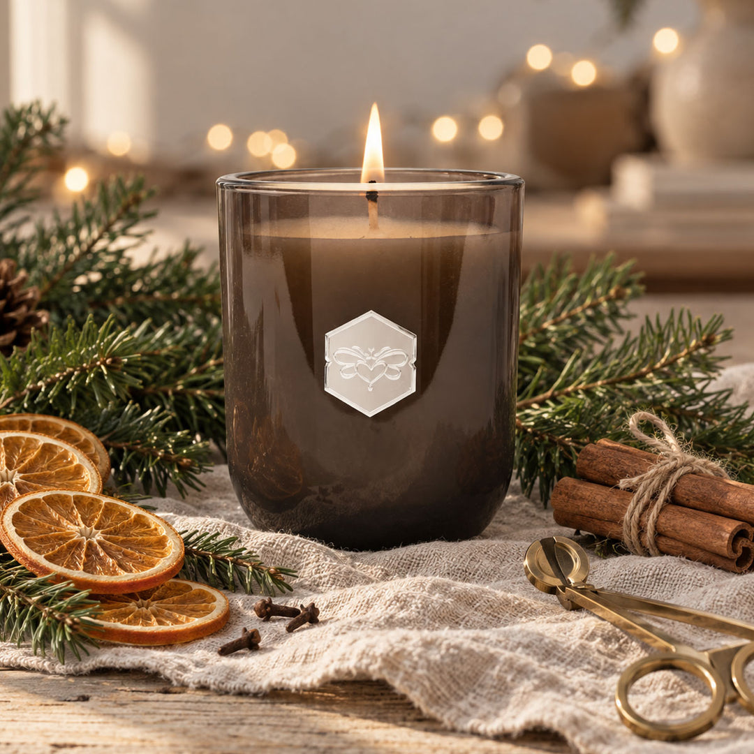 A lit candle in a dark amber glass jar sits on a rustic cloth surrounded by pine branches, dried orange slices, cinnamon sticks, and brass spoons, with warm blurred lights in the background.