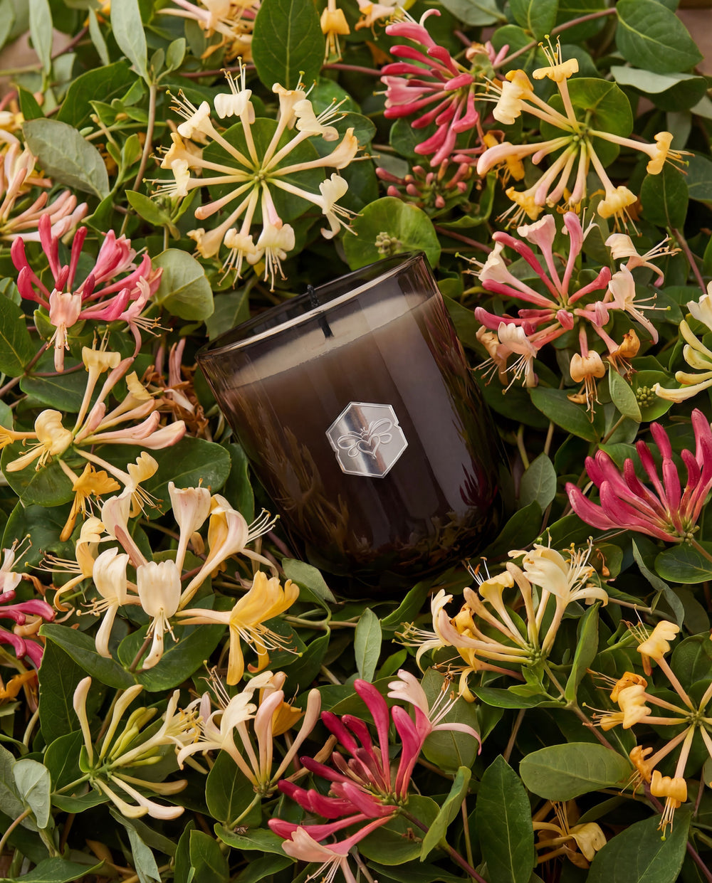 Candle with a brand logo surrounded by pink, yellow, and white flowers on green leaves.
