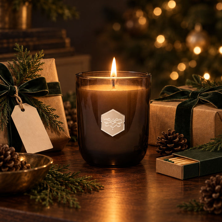 A lit dark amber glass candle with a silver hexagonal logo on the front, styled on a wooden table with wrapped gift boxes, pinecones, evergreen branches, and a blurred Christmas tree in the background.